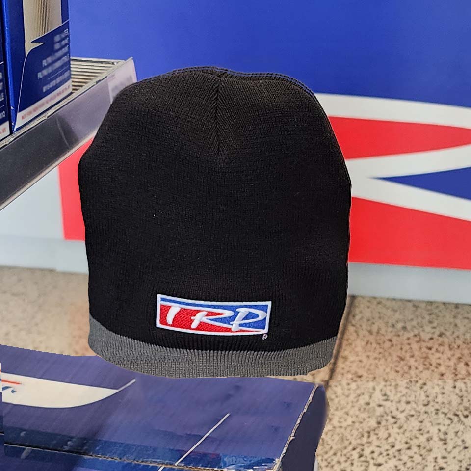 The BEANIE TRP BLACK/GREY, featuring a rectangular red, white, and blue logo patch, is displayed on a blue shelf in a store with a vibrant blue and red background.