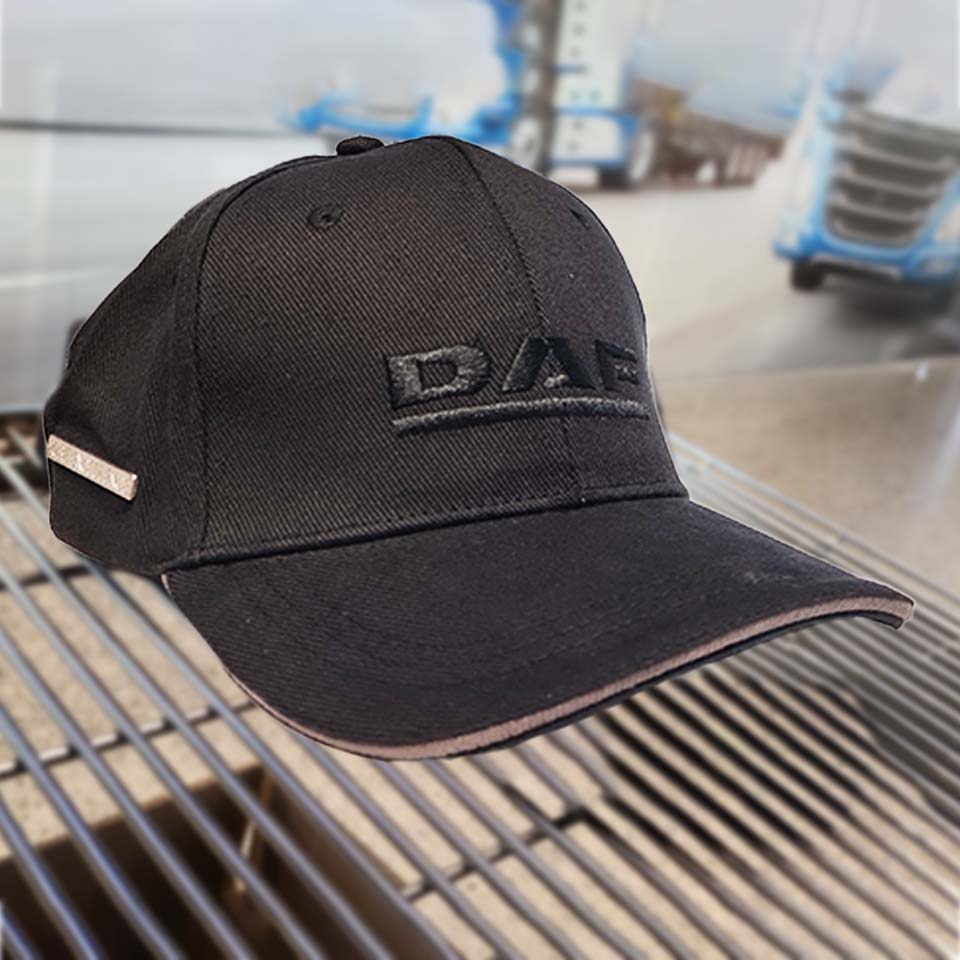 A black baseball cap with DAF embroidered on the front rests on a metal surface, with a blurred background showing blue trucks and an industrial setting.
