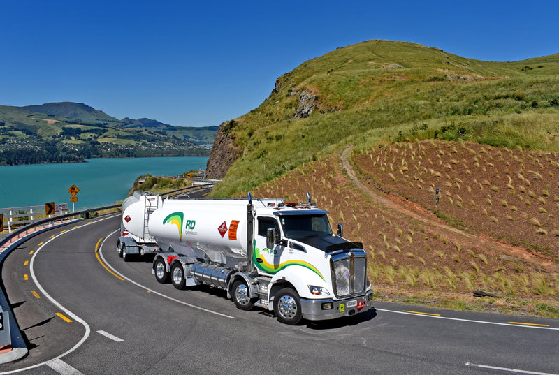 A white fuel tanker truck drives on a winding road beside a blue lake with green hills and mountains in the background under a clear blue sky.