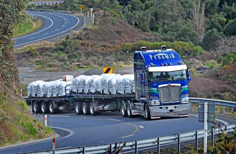 A large blue semi-truck with PYRAMID written on the top hauls a flatbed trailer loaded with white-wrapped cargo, driving on a winding road through a hilly, forested area.