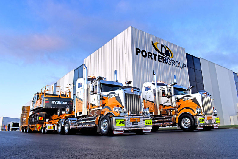 Two large orange and white trucks with “OVERSIZE” signs are parked outside a PORTER GROUP building; one truck is hauling a Hyundai excavator on a flatbed trailer.