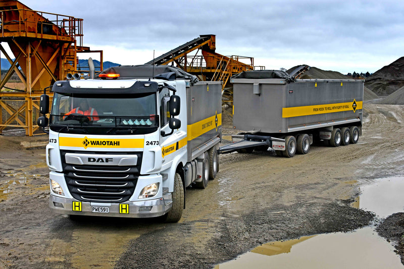 A large Waiotahi-branded DAF dump truck with a trailer is parked at a construction or quarry site with gravel, water puddles, and industrial equipment in the background under a cloudy sky.