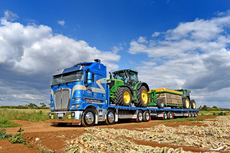 A large blue semi-truck transports a green tractor and yellow farm equipment on a flatbed trailer along a dirt road in a rural field under a partly cloudy sky.