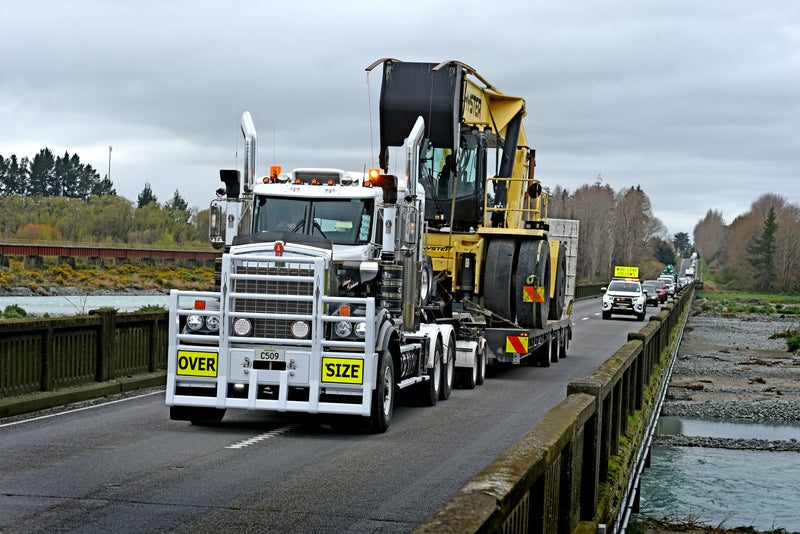 A large white truck marked OVER SIZE transports heavy machinery across a narrow bridge, followed by escort vehicles with warning signs. Trees and a river run alongside the road under a cloudy sky.