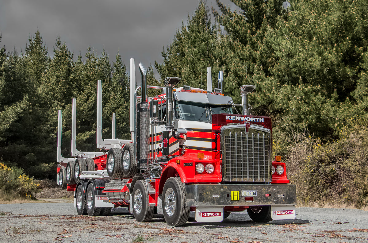 A bright red Kenworth logging truck with empty log bunks is parked on a gravel area with a backdrop of dense pine trees under a cloudy sky.