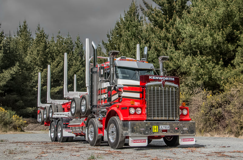 A bright red Kenworth logging truck with empty log bunks is parked on a gravel area with a backdrop of dense pine trees under a cloudy sky.