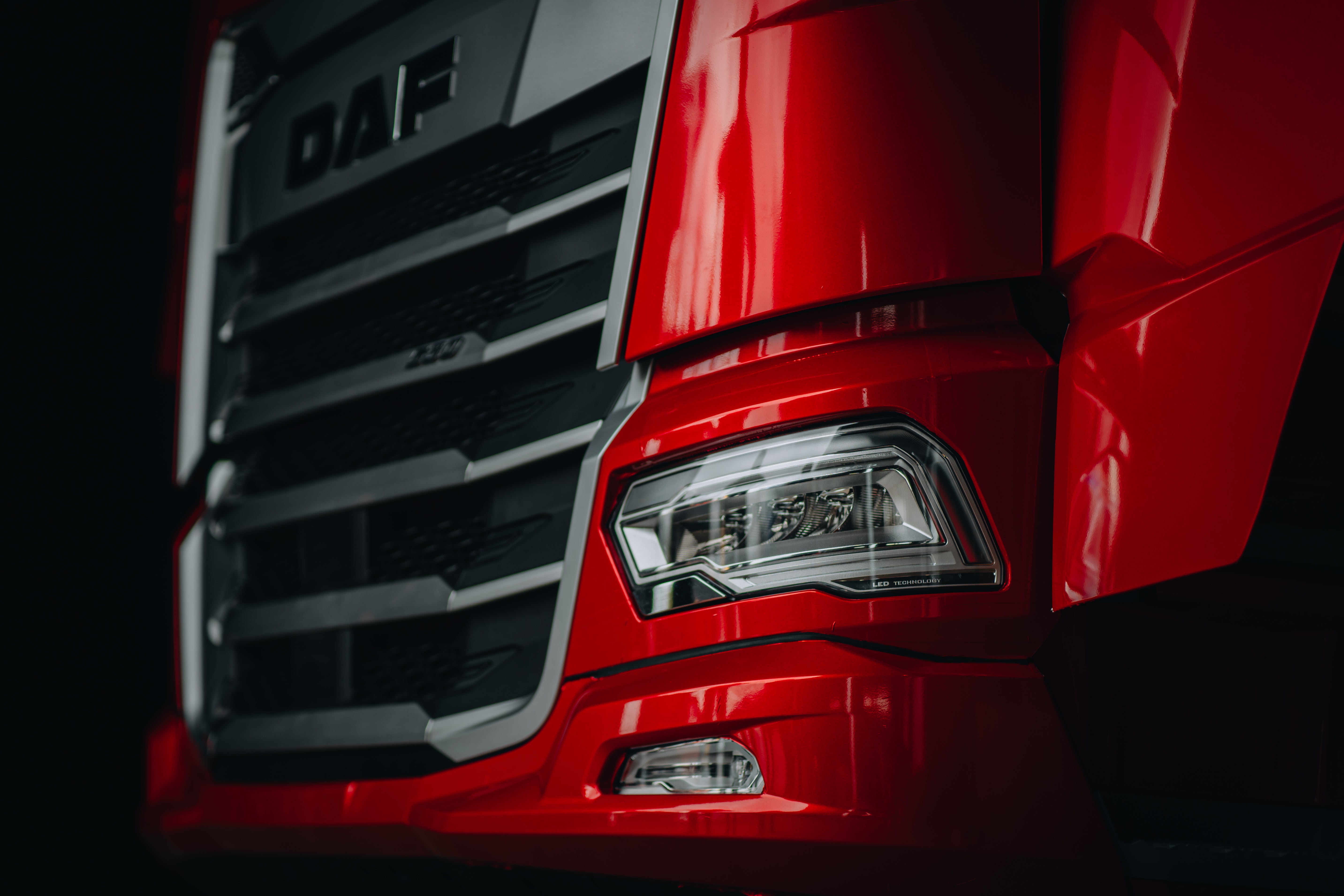 Close-up view of the front corner of a red DAF truck, showing the headlight, grille, and part of the bumper against a dark background.