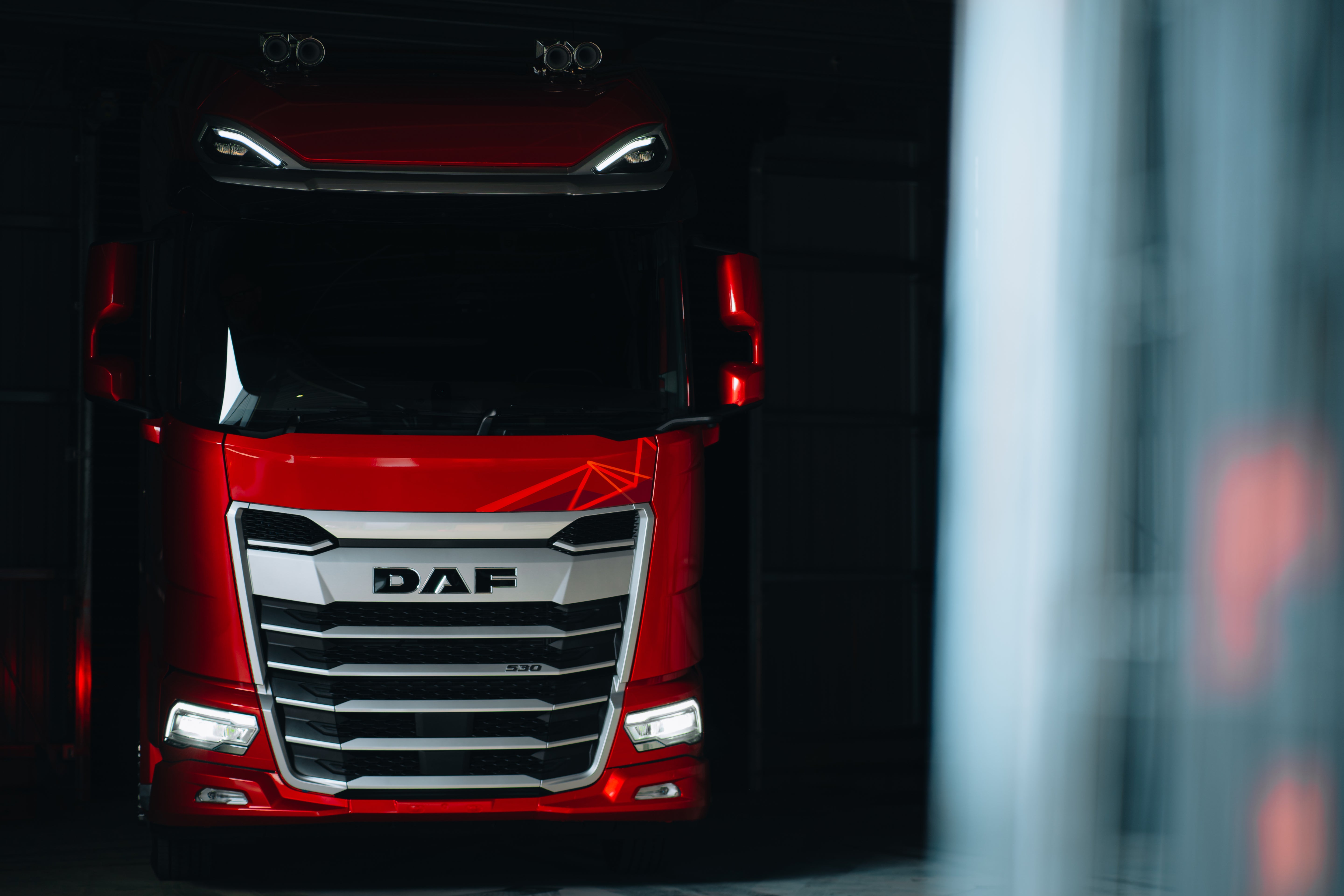 A red DAF truck is parked indoors in a dimly lit space, with its headlights on and front grille prominently displayed.