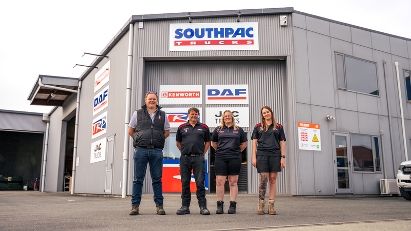 Four people in uniforms stand in front of a Southpac Trucks building, which displays Kenworth and DAF signs. The group is smiling and posing for a photo outside the warehouse-style facility.