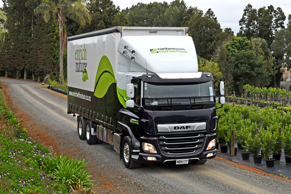 A large DAF truck with Rainbow Park Nurseries branding drives on a gravel road bordered by greenery and potted plants, with tall trees and a garden in the background.