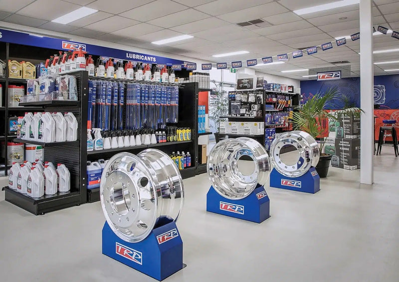 An automotive parts store interior with shiny metal truck rims on display stands, shelves filled with oil containers, windshield wipers, and various car maintenance products in a bright, organized space.