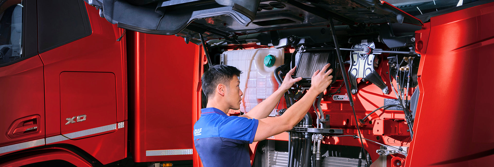 A mechanic in a blue uniform inspects and works on the engine of a large red truck with the hood lifted, revealing the engine components.
