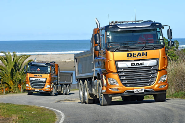 Two large orange DAF dump trucks labeled DEAN drive on a curving road near the coast, with the ocean and sandy beach visible in the background under a clear blue sky.