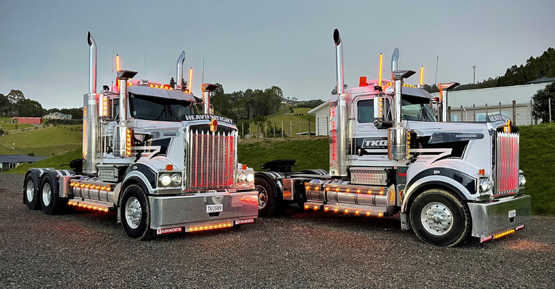Two large, shiny semi-trucks with chrome detailing and bright orange lights are parked side by side on a gravel lot, with grassy hills and buildings visible in the background.