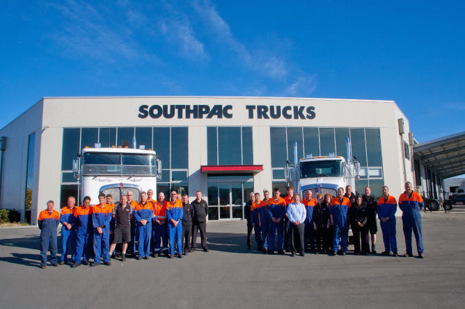 A group of employees in blue and orange uniforms and business attire stand outside a large building labeled SOUTHPAC TRUCKS, with two trucks parked in front under a clear blue sky.