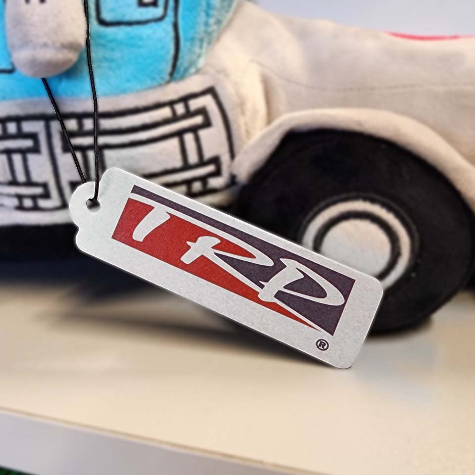 The AIR FRESHENER TRP 85x25mm features a plush truck design and a hangable tag displaying the TRD logo in bold red, white, and blue. The black-string tag doubles as an air freshener with a crisp green apple scent.
