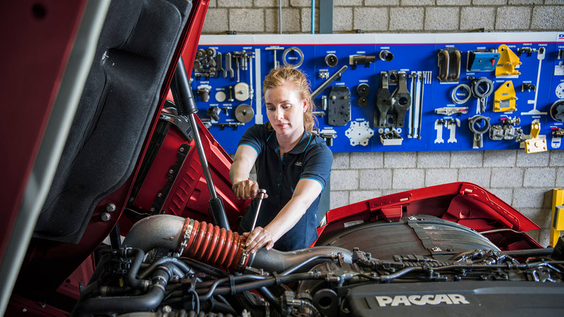 A woman works on the engine of a red truck in a workshop, using a wrench. Behind her, a blue wall-mounted board displays various tools and mechanical parts.