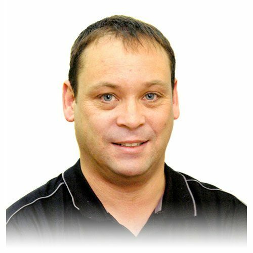 A man with short brown hair and blue eyes is smiling slightly. He is wearing a black collared shirt with white piping, and the background is plain white.