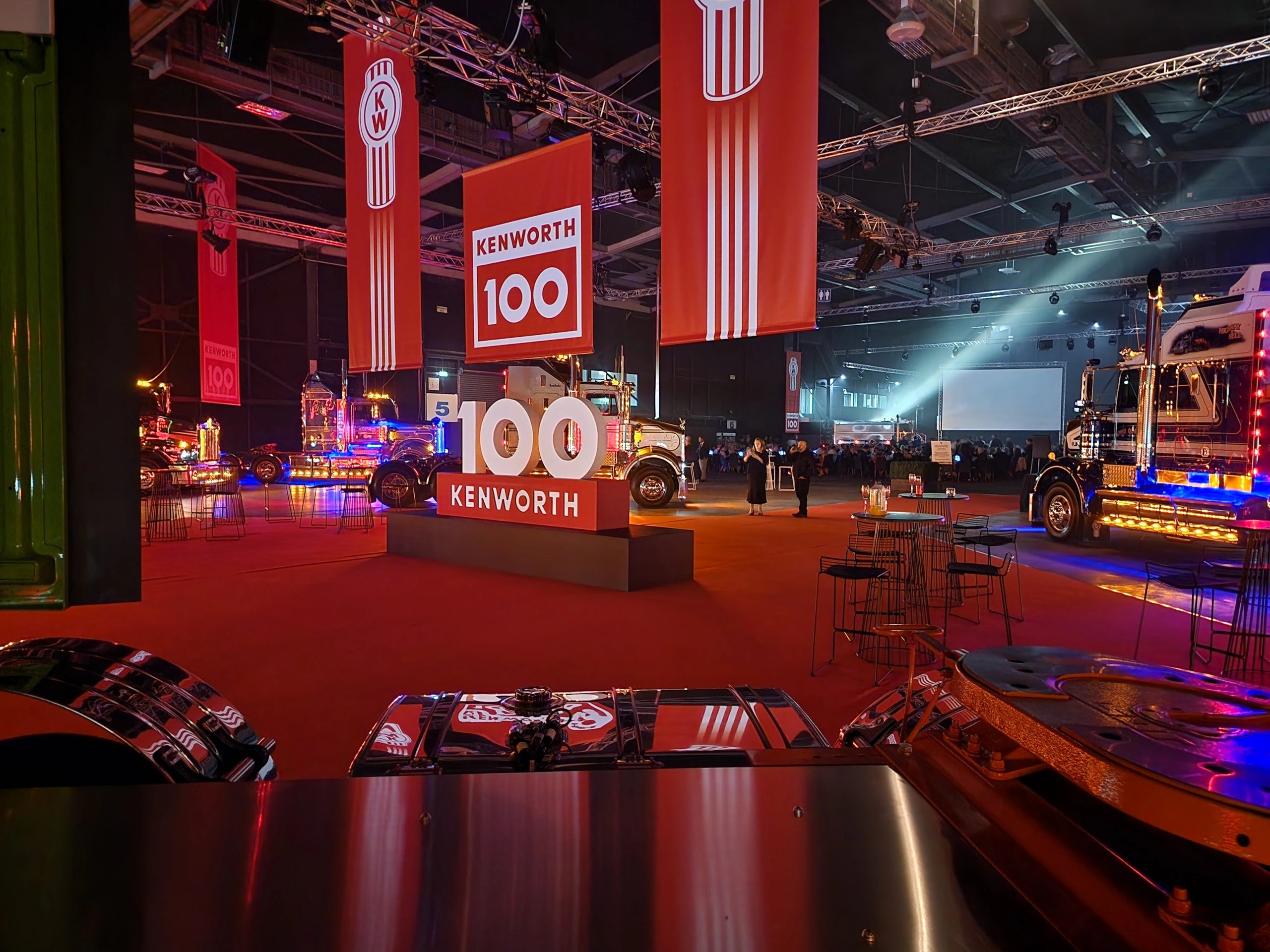 A large indoor event celebrates Kenworths 100th anniversary, featuring illuminated truck displays, Kenworth 100 signs, banners, spotlights, and tables with chairs on a red carpet. People are visible in the background.