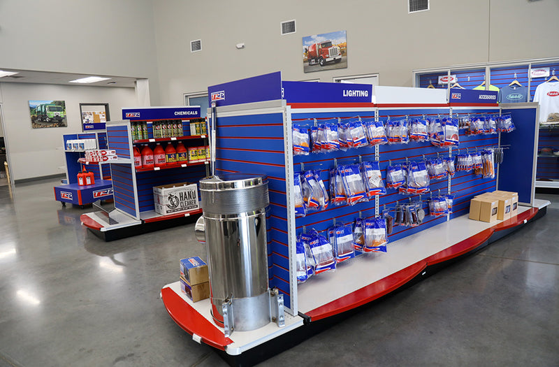 A clean store interior with shelves displaying truck parts and accessories, including lighting components, chemicals, filters, boxes, and branded merchandise on the walls; the floor is polished concrete.