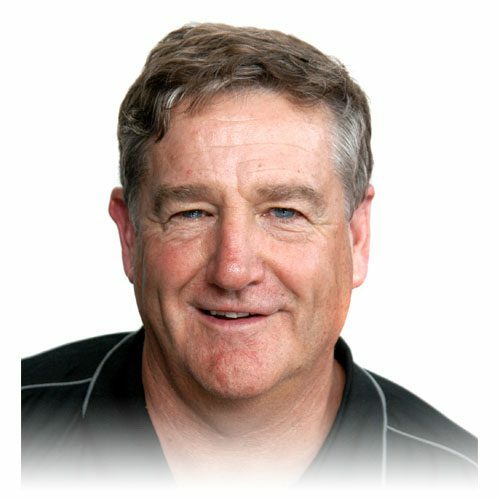 A middle-aged man with short, light brown hair and blue eyes, smiling. He is wearing a black collared shirt with white piping and is pictured against a white background.