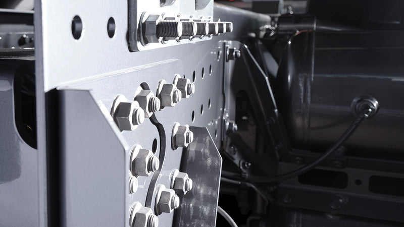 Close-up of a metallic machine or vehicle component, showing multiple bolts, nuts, and plates attached to a grey metal frame, with cables and additional metal parts in the background.