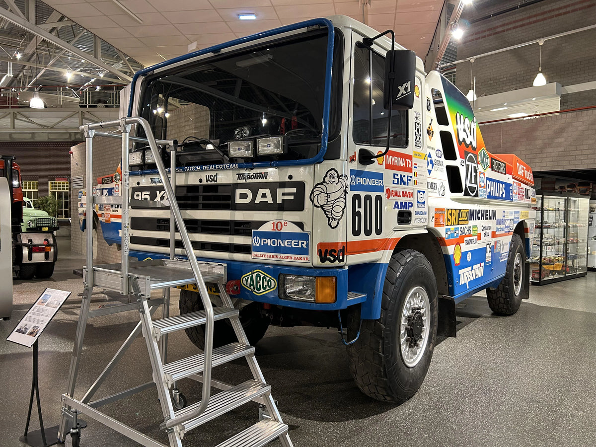 A large, colorful DAF racing truck covered in sponsor logos is displayed indoors, with a metal staircase leading to its cab. The truck is part of an exhibit, with informational signs nearby and other vehicles in the background.